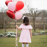 Child in a pink dress holding red and white heart balloons in a park.