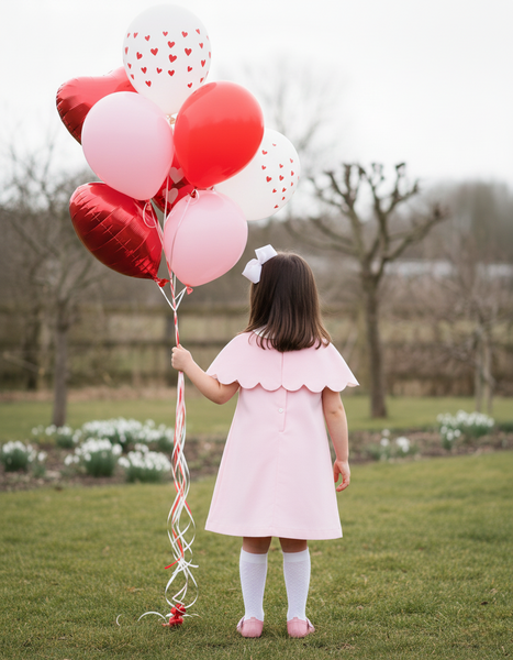 Child in a pink dress holding red and white heart balloons in a park.