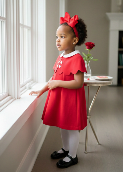 Young girl in a red dress standing by a window with a rose on a small table.