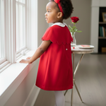 Young girl in a red dress standing by a window in a bright room.