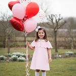 Young girl in a pink dress holding red and white heart-shaped balloons in a park.