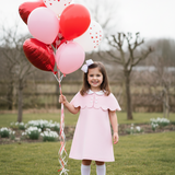 Young girl in a pink dress holding red and white heart-shaped balloons in a park.