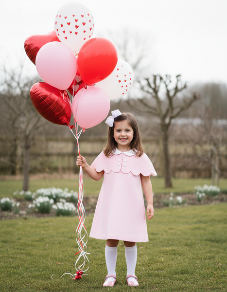 Young girl in a pink dress holding red and white heart-shaped balloons in a park.