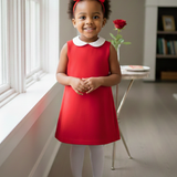 Young girl in a red dress with a large bow standing in a room with a window and bookshelf.